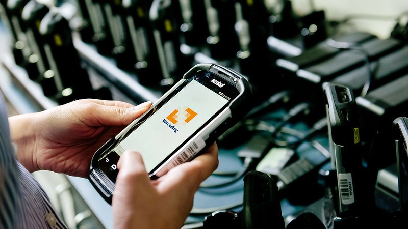 Barcode scanner with barcoding logo on it in front of station with many scanners being charged